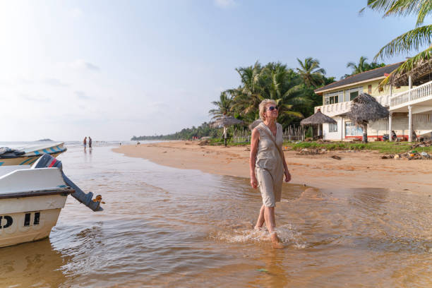 . A woman in a flowing dress walks on the beach beside a boat, embracing the peaceful atmosphere of the coastal landscape.