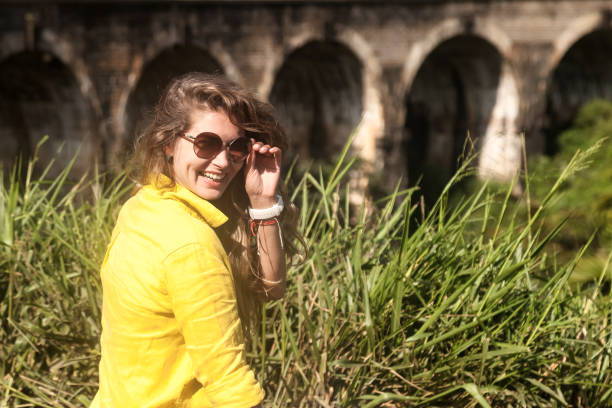 A woman wearing a yellow shirt and sunglasses stands confidently in front of a bridge, enjoying the sunny day.