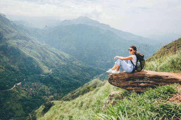 A woman sits on a log, surrounded by majestic mountains, enjoying the serene natural landscape.