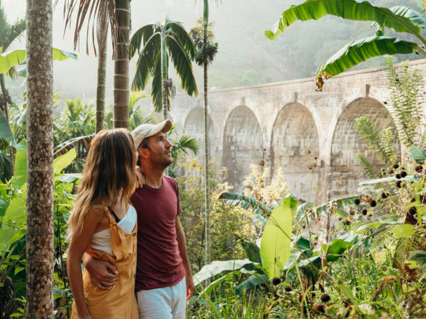 A couple gazes at a bridge in a lush tropical jungle, surrounded by vibrant greenery and exotic plants.