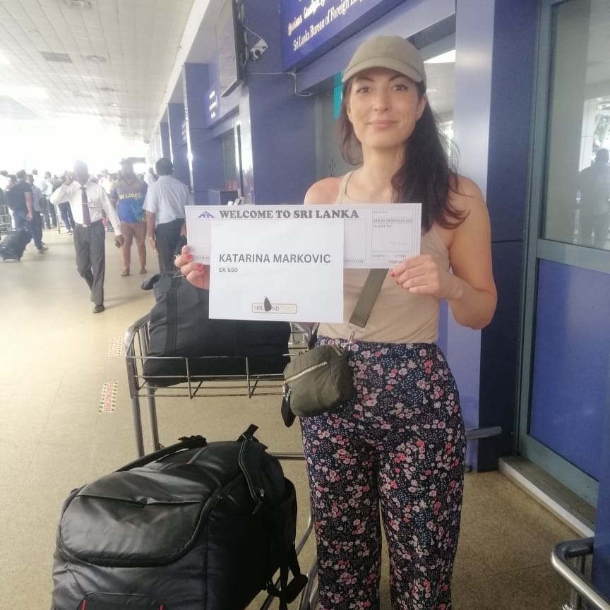 A woman stands at an airport, holding a sign that reads "Airport Transfer Sri Lanka," ready to assist travelers.