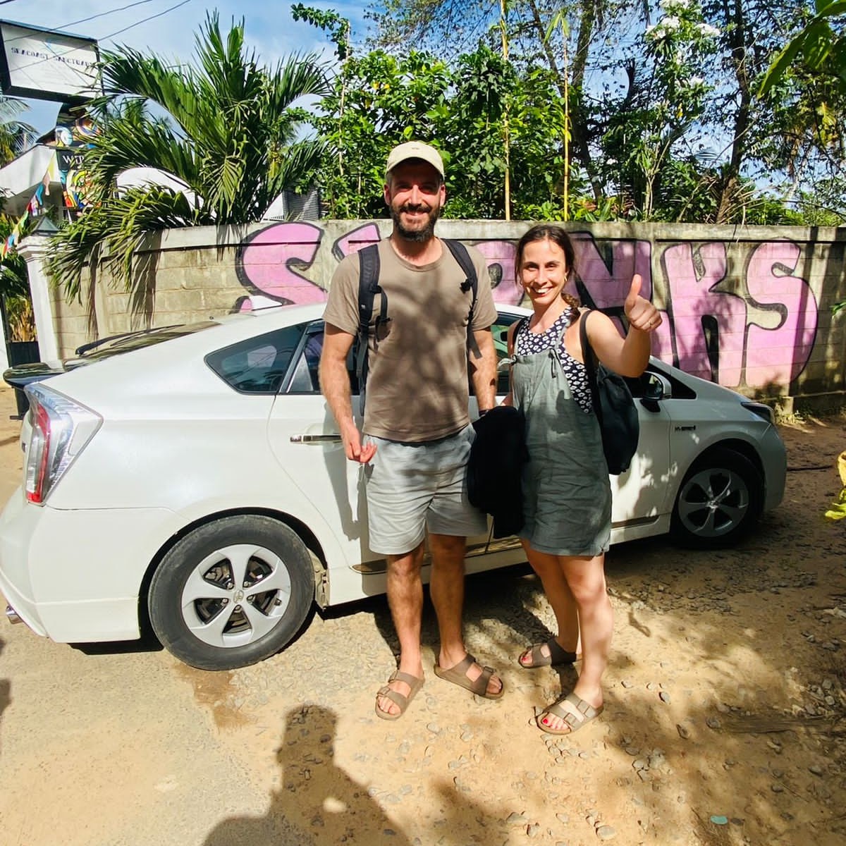 A man and woman stand beside a white taxi, ready to provide a taxi service to their passengers.