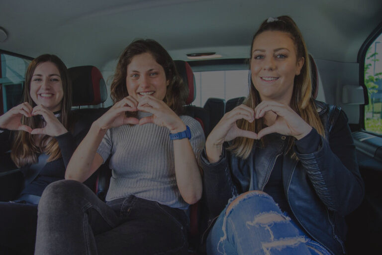 Three women in a car forming a heart shape with their hands, showcasing friendship and joy in a cozy setting.